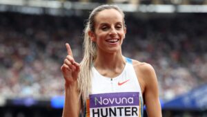 Georgia Hunter Bell standing with her finger in the air smiling for the cameras in the stadium in her running kit and her blonde hair in a pony tail.